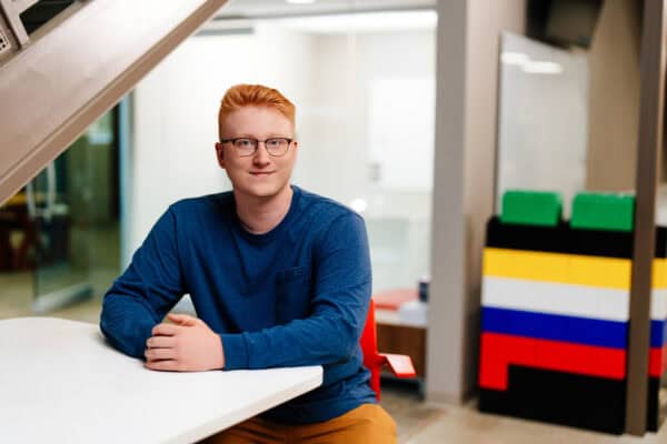 A young man with red hair and glasses is seated at a white table in a modern office environment. He is wearing a blue long-sleeve shirt and has a friendly expression. In the background, there is a colorful structure made of large building blocks, and the office has a clean, contemporary design with glass partitions and light-colored walls.