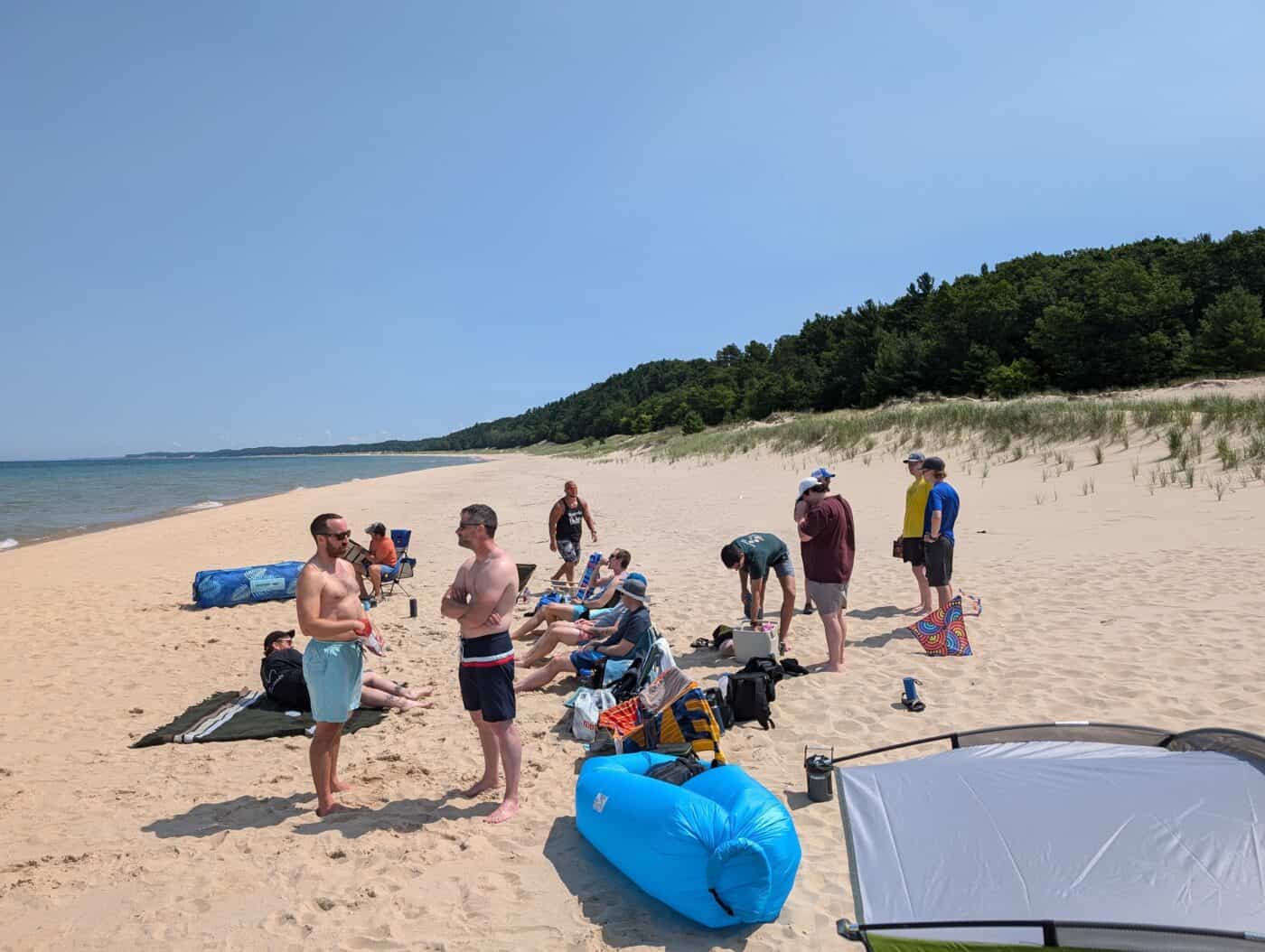 The image shows a group of people enjoying a sunny day at the beach. The beach is sandy and stretches out towards the horizon, bordered by calm, blue water on one side and lush green trees on the other. The sky is clear with no clouds, indicating a perfect summer day. Several individuals are gathered, some standing and talking while others are sitting on beach chairs or towels. A few people are seen preparing items or relaxing. In the foreground, there is an inflatable blue lounger and some beach gear, including a colorful beach umbrella and a cooler. The overall scene conveys a lively and relaxed atmosphere of friends or family enjoying a day out by the water.