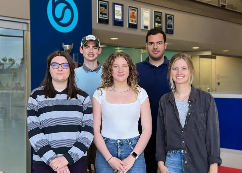 This image shows a group of five interns standing together in a corporate office setting. They are positioned in two rows, with three people in the front and two in the back. The interns are casually dressed, with a mix of smiles and neutral expressions. The background includes a blue wall with a logo and several award plaques displayed above. The overall atmosphere appears professional and welcoming.