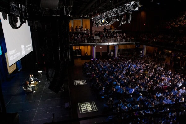 This image shows a large indoor event from Tech Week Grand Rapids 2023. The venue is filled with a large audience seated in front of a stage where two speakers are engaged in a discussion. The stage has a big screen displaying event information, including a prompt for submitting Q&A questions and a QR code. The lighting is focused on the stage, while the audience is dimly lit. There is also "Tech Week Grand Rapids" branding projected onto the floor near the stage. The overall atmosphere suggests a professional tech conference or summit, with a significant turnout of attendees.