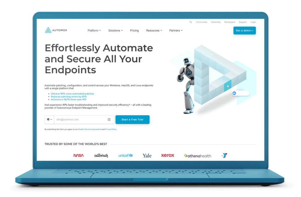 A laptop displaying the Automox website homepage on a white background. The screen features a headline that reads 'Effortlessly Automate and Secure All Your Endpoints,' along with a futuristic robot graphic and a call-to-action button for starting a free trial. Below, logos of well-known companies such as NASA, Xerox, and Yale are displayed as trusted clients.