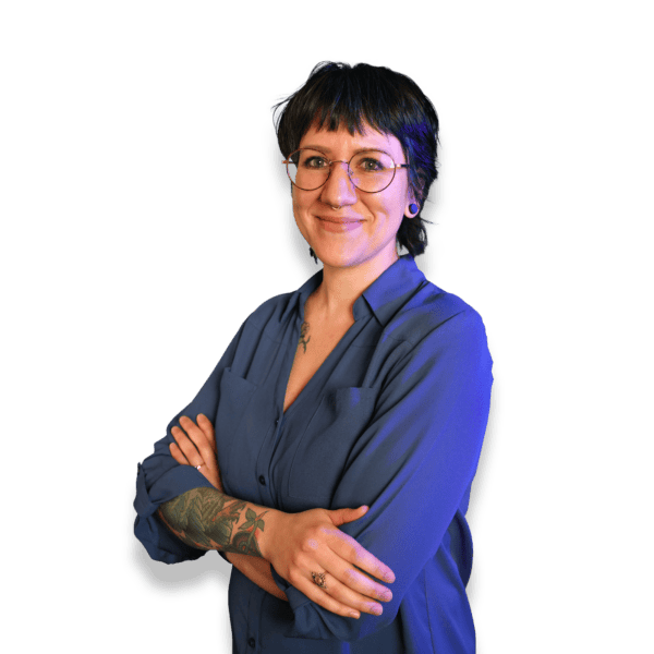 Person with short dark hair, wearing glasses and a blue shirt, standing confidently with arms crossed against a black background, showcasing visible tattoos on their forearm.